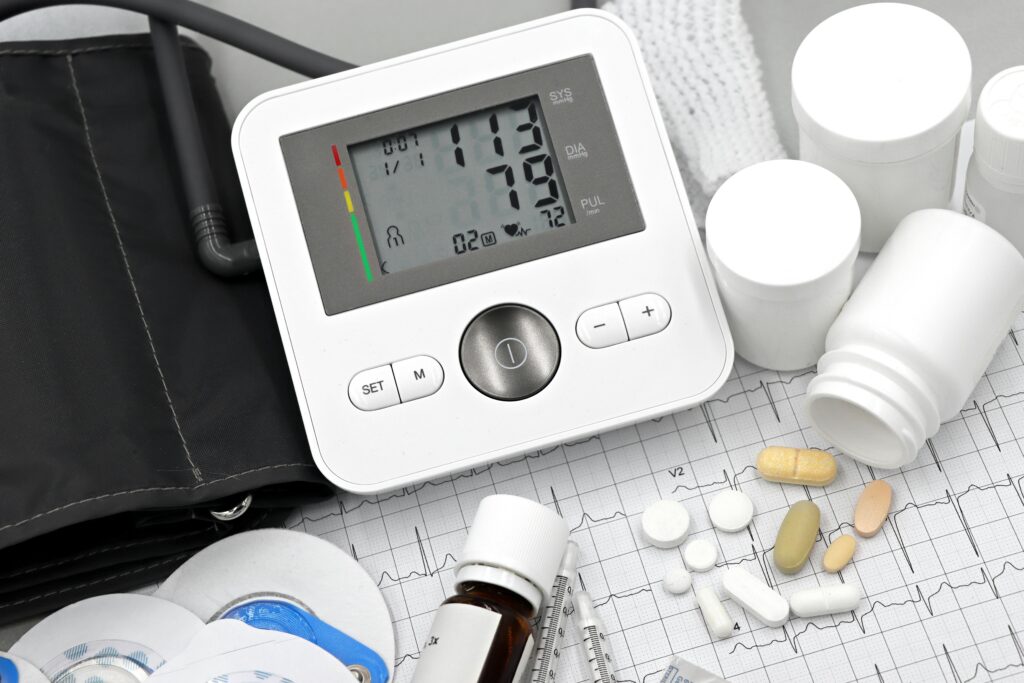 Digital blood pressure monitor with cuff on an ECG printout, surrounded by pills, syringes, and medicine bottles.